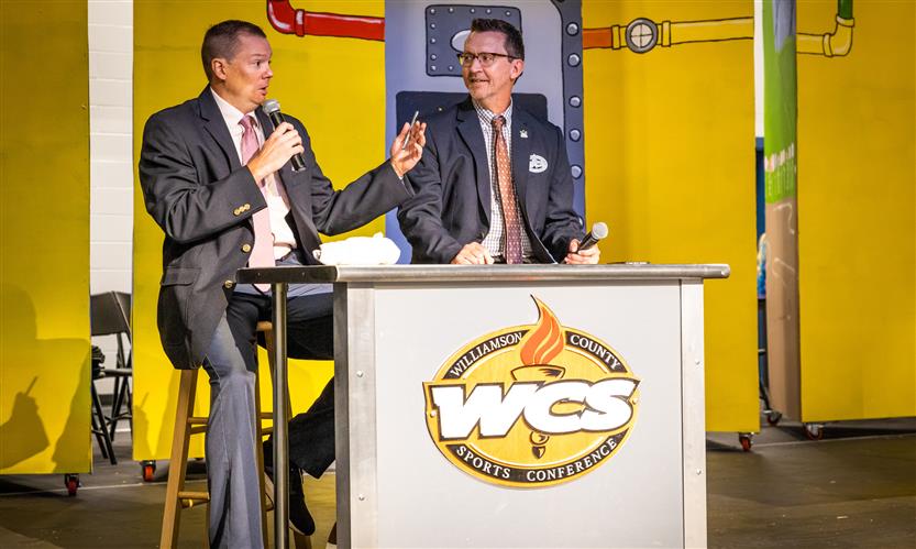 Two men sit at a desk onstage and speak